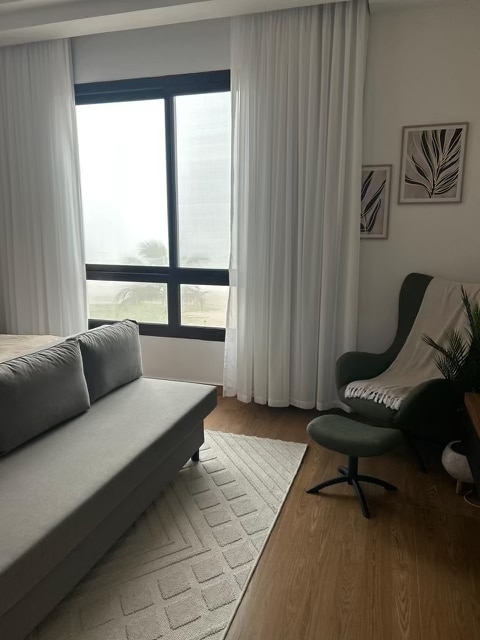 A cozy corner of the studio features a comfortable sofa and a stylish green armchair positioned near a large window. Soft, sheer curtains allow natural light to filter in, creating a bright and airy environment. A patterned area rug adds warmth to the space.