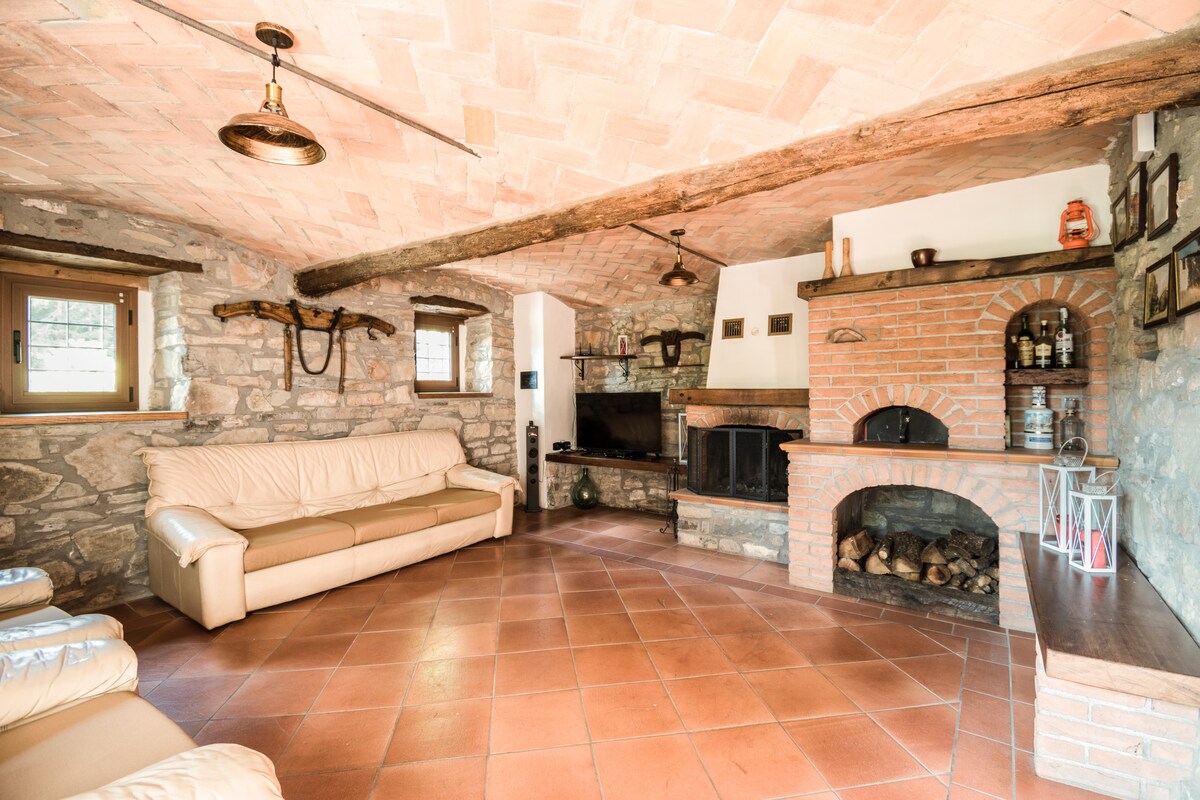 A spacious living area features exposed wooden beams and a warm terracotta tile floor. A stone fireplace with a brick surround holds firewood, and a television sits atop a rustic console. Comfortable seating is provided by a light-colored leather sofa, enhancing the cozy atmosphere.