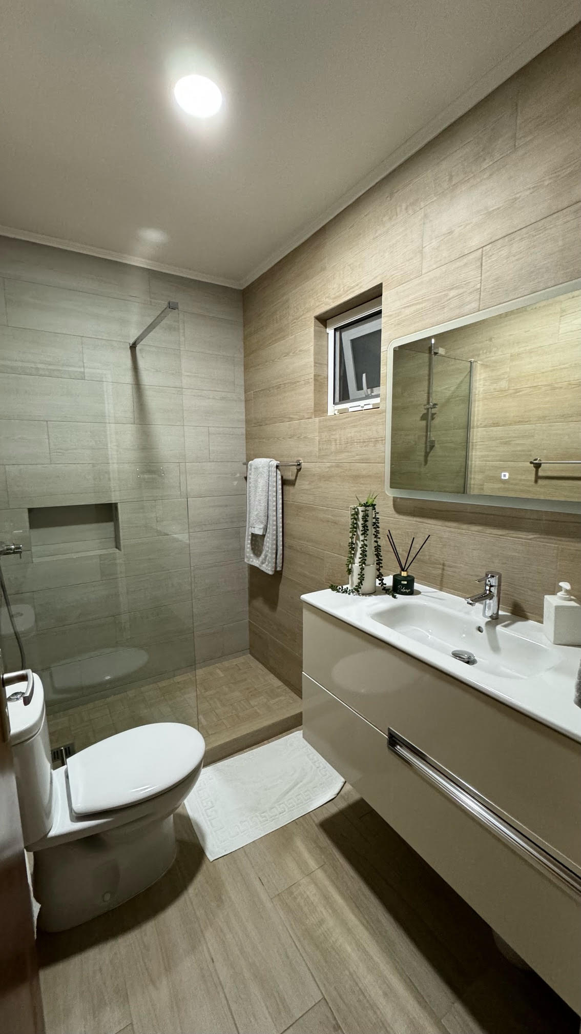 A modern bathroom features a glass shower enclosure and a compact toilet. A sleek vanity with a wide sink is complemented by a large mirror above it. Neutral-toned tiles cover the walls and floor, while a small window allows for natural light.