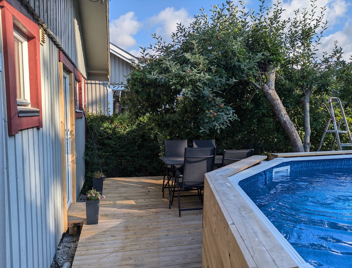 A wooden deck features a round pool and a dining area with a table and six chairs. Surrounding greenery provides shade and privacy. The area is accessed through a door from the house, creating a seamless transition to outdoor relaxation.