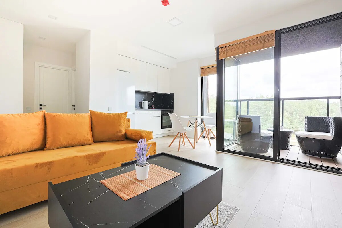 A contemporary living area features a vibrant gold sofa and a black coffee table with a decorative plant. Light enters through large glass doors, leading to a balcony. A dining space with white chairs and a modern kitchen area is also visible in this well-lit room.