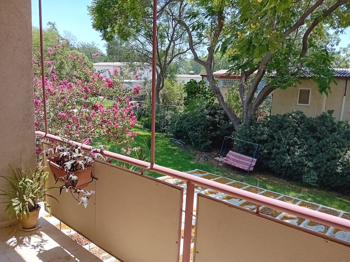 A small balcony features a railing adorned with potted plants, offering views of a lush green landscape. Colorful flowers bloom nearby, complemented by a swing in the garden. Tall trees provide shade, creating a serene outdoor setting for relaxation.