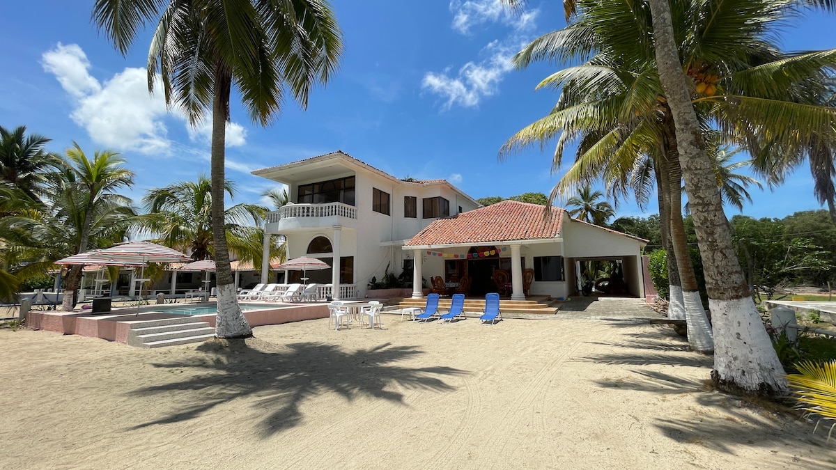 A spacious two-story villa is positioned near the beach, surrounded by palm trees. A private pool is visible alongside lounge chairs and umbrellas on the sandy shore. The entrance features steps leading to a covered patio area, providing access to the indoor living spaces.