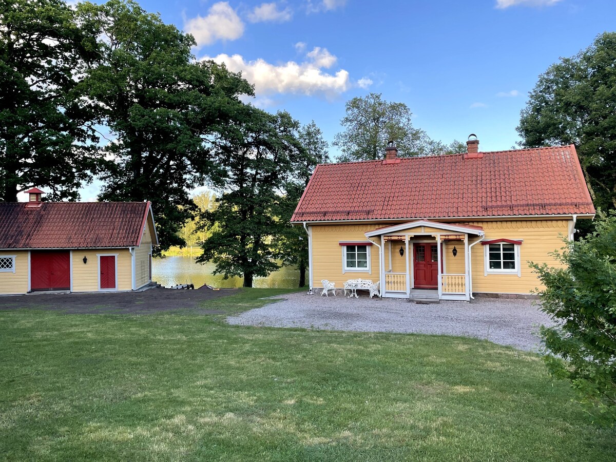 A charming yellow house is set against a backdrop of lush green trees and a tranquil lake. The property features a spacious lawn, with a gravel area leading to a cozy sitting arrangement in front of the home. A separate red structure is visible to the left.