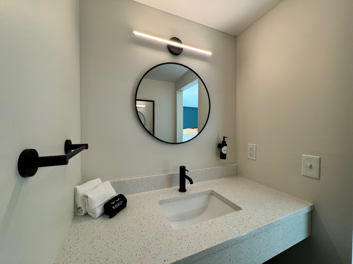 A modern bathroom sink area is shown, featuring a white countertop with a smooth finish and a circular mirror above. A sleek black faucet and wall-mounted soap dispenser contrast with the light-colored walls. Clean white towels are neatly arranged on the countertop.