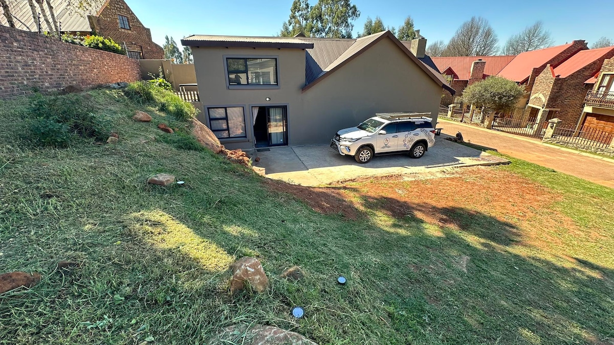 The exterior view of the recently renovated holiday home displays a modern two-story structure with a large driveway. A vehicle is parked beside the house on a concrete pad, surrounded by a grassy area and rocky landscaping, set against a clear blue sky.