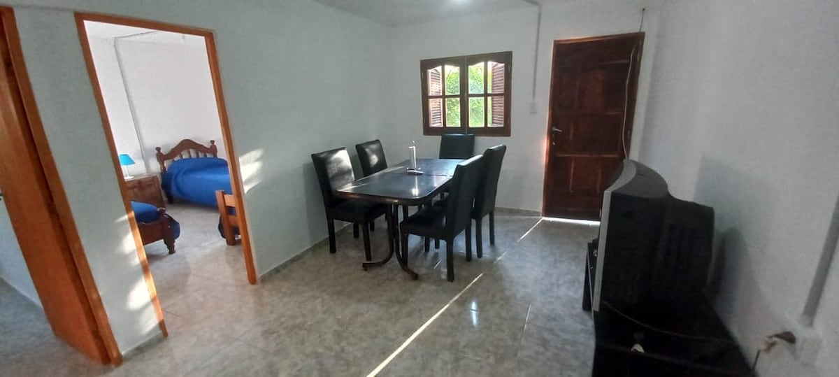 A dining area is shown with a round table surrounded by six black chairs. Natural light enters through a window, illuminating the space. A wooden door leads outside, while a glimpse of a separate sleeping area is visible in the background.