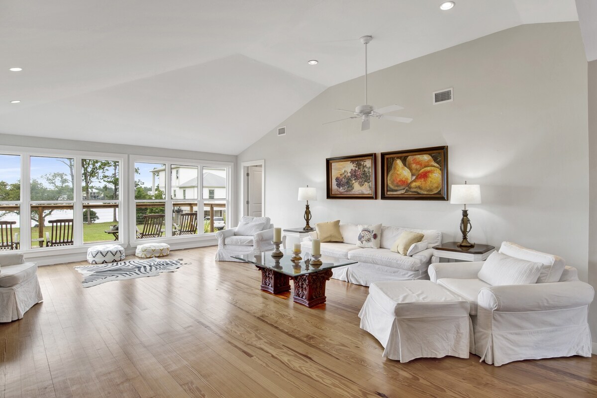 A spacious living area features light-colored upholstery and wooden flooring. Large windows provide natural light and views of the outdoor space. Decorative artwork adorns the walls, while a coffee table serves as a central focal point surrounded by comfortable seating.