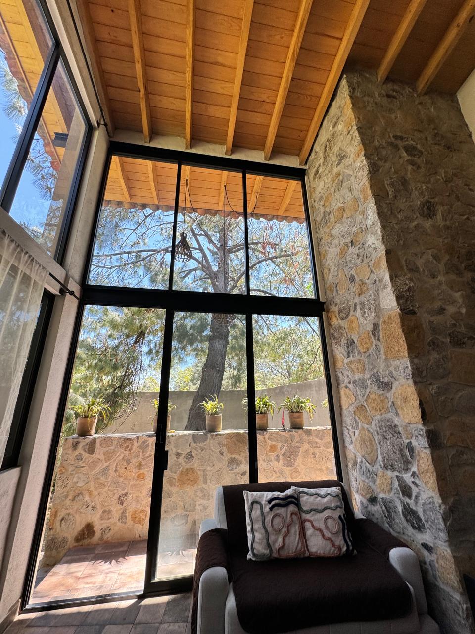 A sunlit corner is framed by large glass windows, showcasing a view of trees and potted plants outside. A cozy seating area features a dark sofa adorned with a decorative pillow, complemented by a natural stone wall that provides warmth to the space.