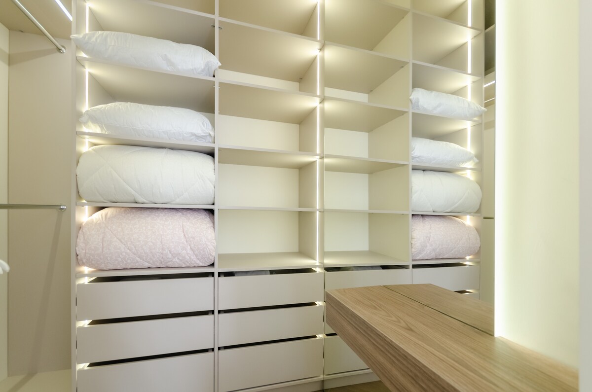 A modern wardrobe display showcases neatly arranged shelves filled with pillows and bedding. Soft, subtle lighting highlights the items, enhancing visibility within the space. A wooden dressing table is positioned in front, providing a functional area for personal items.