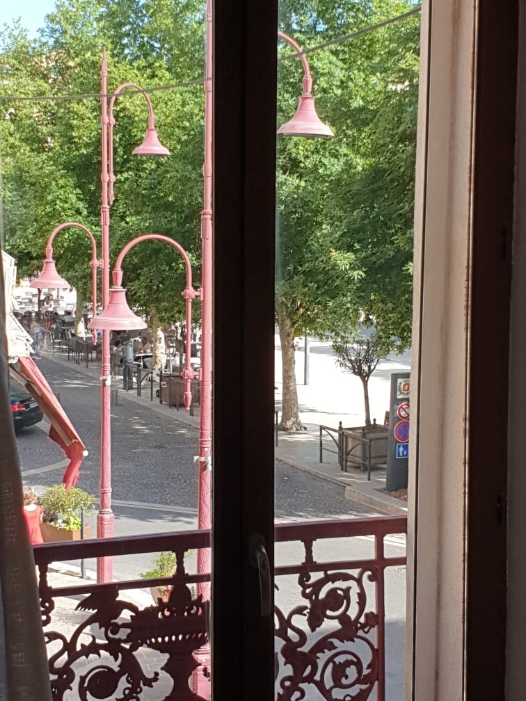 A view from the balcony depicts vibrant street lamps in pink, surrounded by green trees. A tree-lined plaza is visible in the distance, along with outdoor seating areas. Natural light filters through the half-open window, showcasing the architectural details of the balcony railing.
