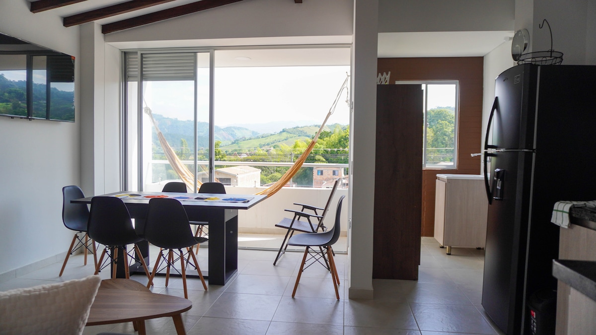 A light-filled living area features a dining table surrounded by chairs, with a cozy hammock visible through expansive glass doors. The view includes rolling hills in the distance. A sleek refrigerator and light wooden cabinetry is positioned nearby, creating a modern yet inviting space.
