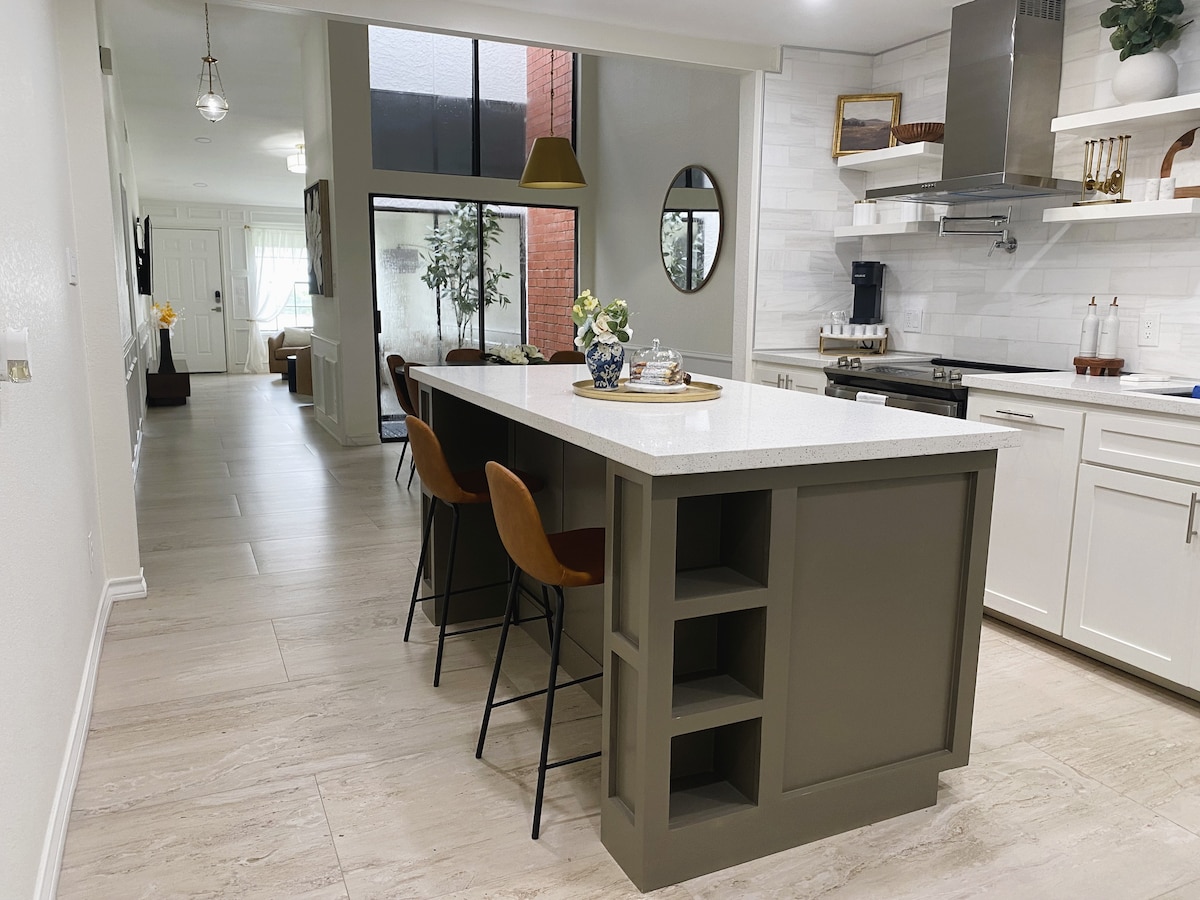A modern kitchen with a central island includes four high-backed stools. Sleek cabinetry and a stainless steel appliance setup enhance functionality. Natural light fills the space through large windows, creating a bright and inviting atmosphere.