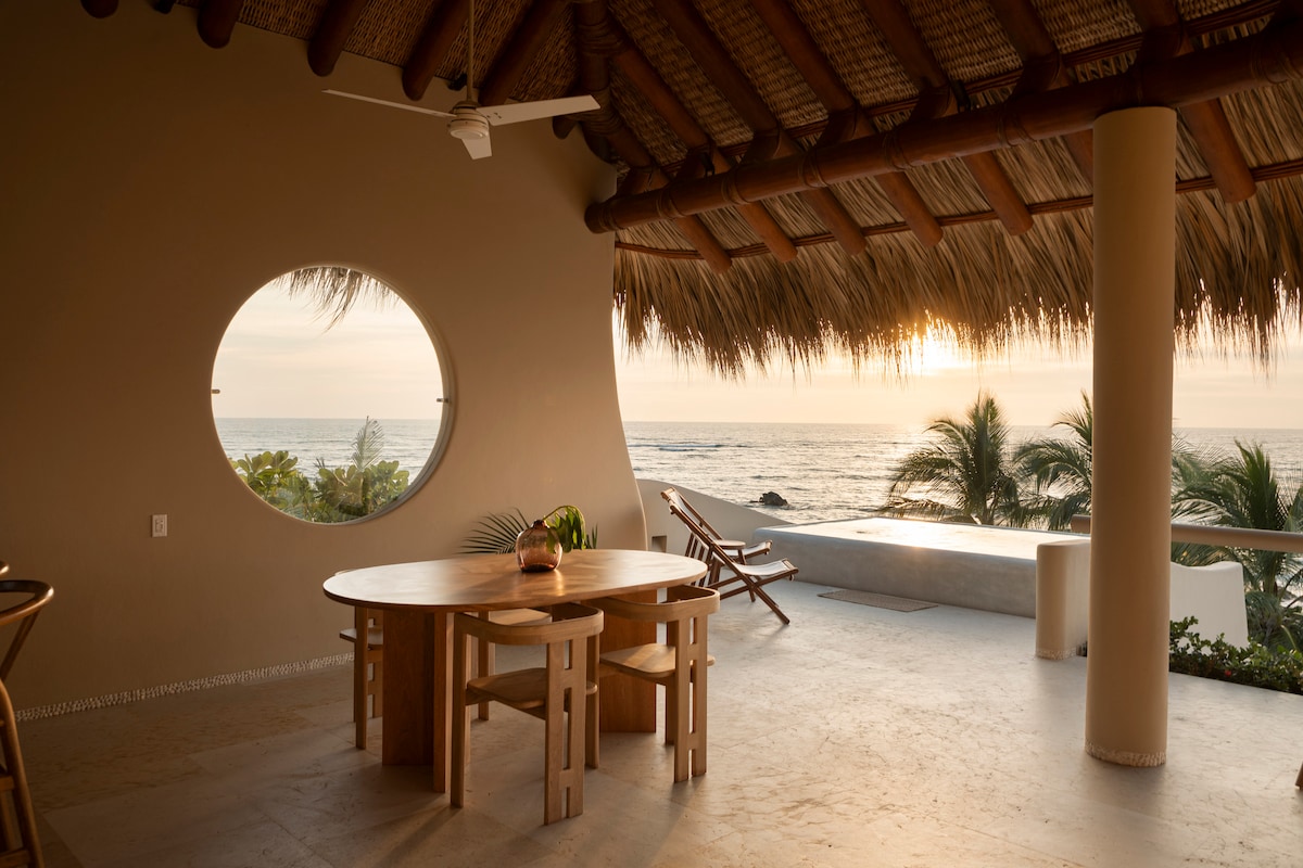 An open dining area is captured with a round wooden table complemented by several chairs. A large circular window frames a view of the ocean, while the soft glow of sunset illuminates the space, enhancing the natural decor with palm trees visible in the background.