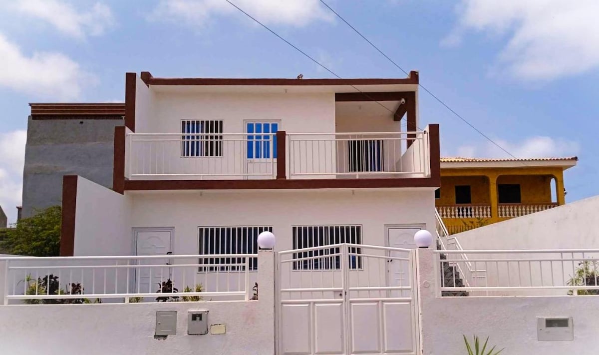 The exterior of a two-story building features a simple yet modern design, with two sets of balcony windows on the upper level. A white fence surrounds the property, complemented by minimal landscaping near the entrance. The sky is bright and partly cloudy, adding to the inviting atmosphere.