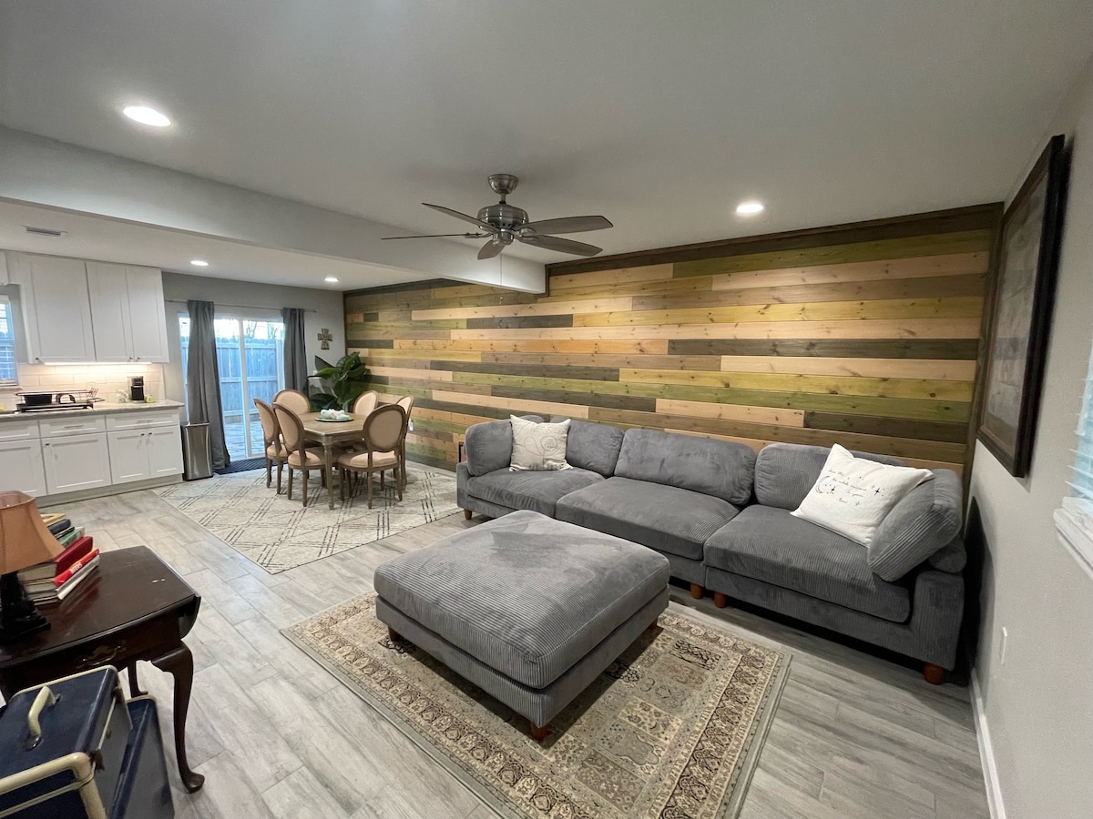 A spacious living area features a large, plush sectional sofa and a coordinating ottoman. A circular dining table surrounded by four chairs is positioned nearby. Natural light enters through sliding glass doors, illuminating the wooden accent wall and tiled flooring.