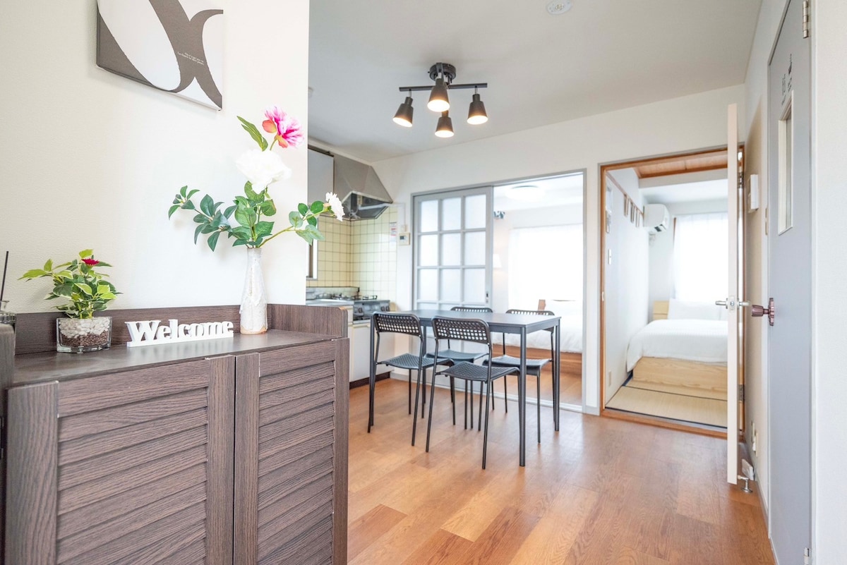An open living area showcases a modern dining table with four black chairs, set against a wooden floor. A kitchenette is visible to the left, with a small decorative plant and a welcome sign on the side table. Natural light fills the space through a window leading to the bedroom.