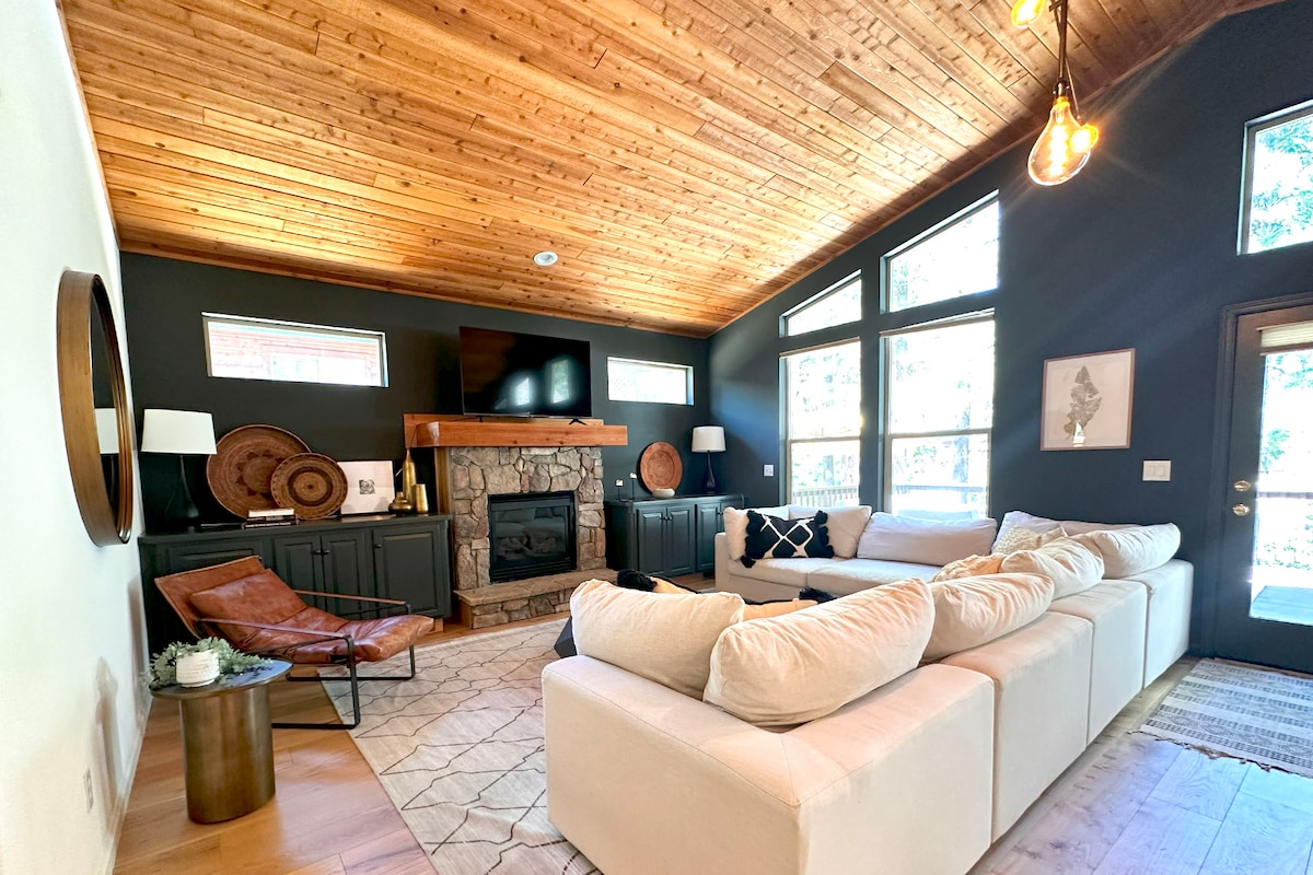 A spacious living area is highlighted by a vaulted wooden ceiling and large windows, allowing natural light to fill the room. A comfortable sectional sofa and a leather lounge chair are arranged around a stone fireplace, with a television mounted above the mantel.