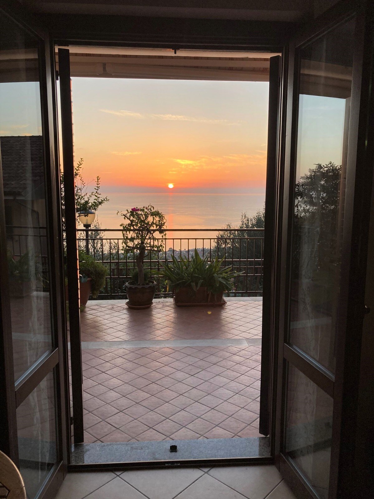 A doorway with glass doors opens onto a terrace, where potted plants line the railing. The view captures a sunset over the calm sea, painting the sky in shades of orange and pink, creating a peaceful ambiance at twilight.