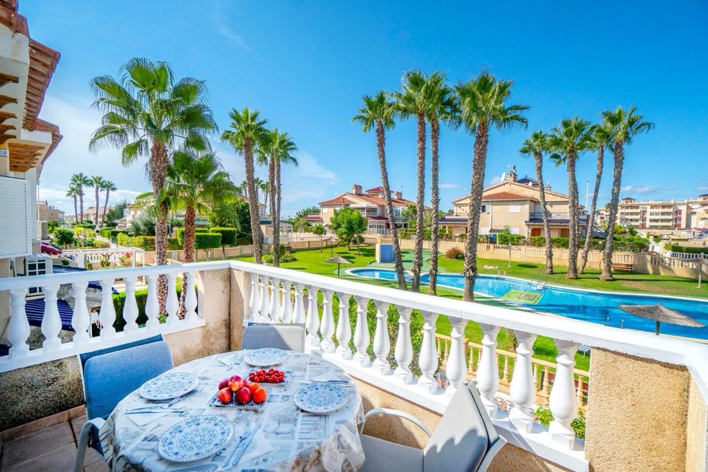 A spacious outdoor terrace is furnished with a dining table set for six, complete with a decorative plate of fruit. Sunlight bathes the area, while palm trees and a large swimming pool are visible in the background, enhancing the inviting outdoor setting.