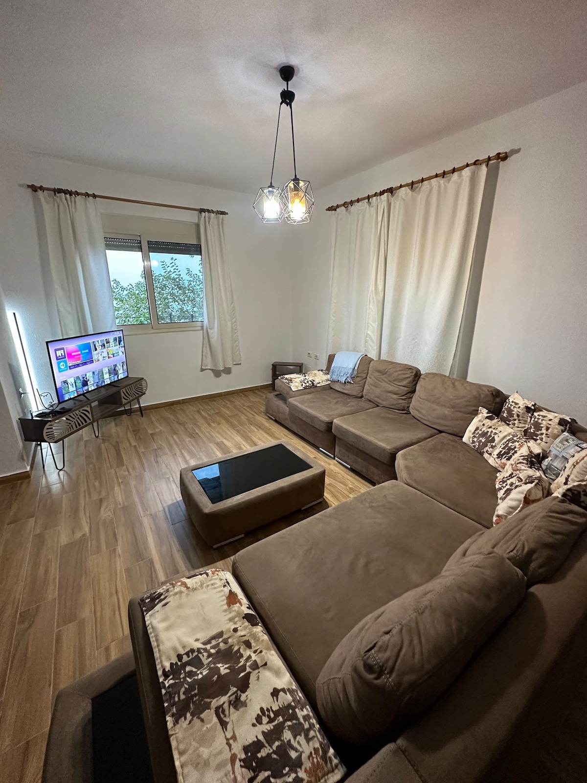 A cozy living area features a large sectional sofa adorned with decorative pillows. A coffee table occupies the center, while a TV sits on a stylish stand. Natural light enters through a window, with sheer curtains framing the view.