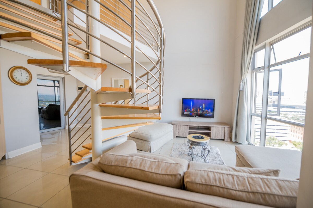 An open-plan living area features a spiral staircase leading to an upper level. A light-toned sofa surrounds a circular coffee table, with a television mounted on the wall opposite. Large windows allow natural light to fill the space, offering views of the city.