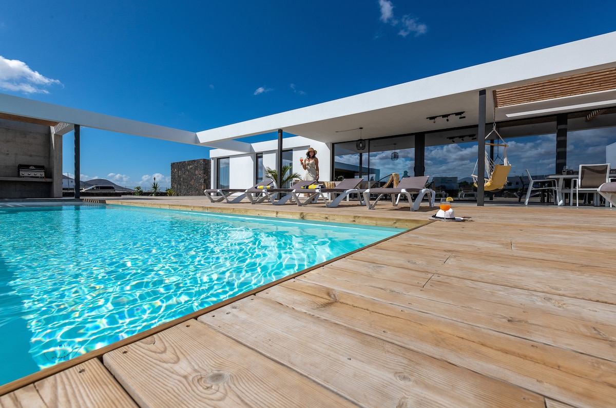 A modern pool area is presented with a wooden deck surrounding a tranquil blue pool. Sun loungers are arranged along the deck, and an outdoor dining area is visible nearby, framed by large glass doors leading into the villa.