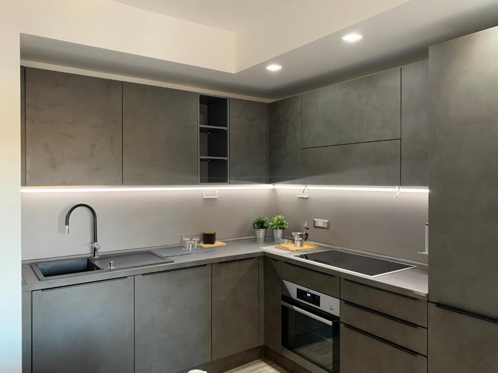A modern kitchen is presented with sleek gray cabinetry and minimalist design. Under-cabinet lighting illuminates the space, highlighting a sink, a stovetop, and an oven. Open shelves are arranged for decorative plants, while a wooden cutting board and glassware are neatly placed on the counter.