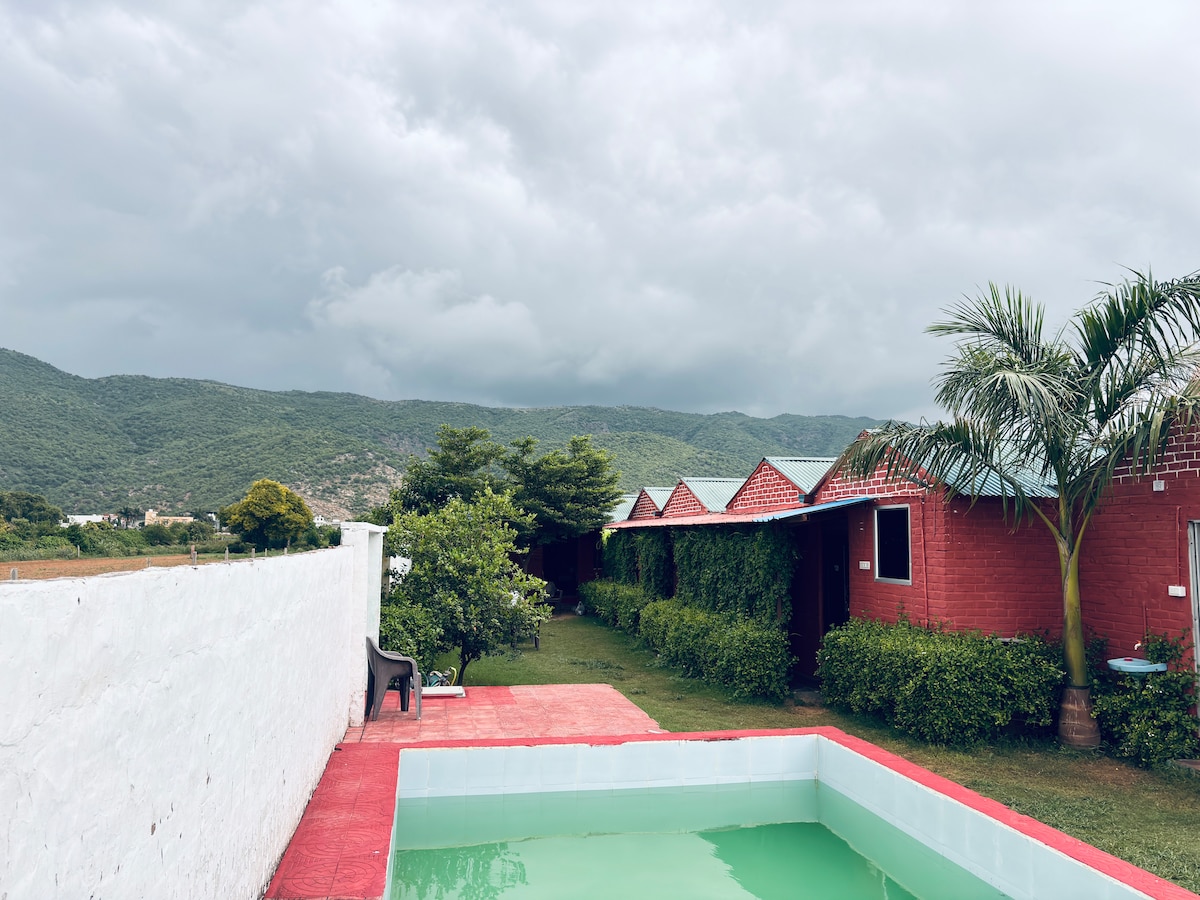 A serene view of the property features a bright red pathway leading to cozy cottages surrounded by greenery. A small pool is visible in the foreground, reflecting the cloudy sky, while rolling hills provide a tranquil backdrop.