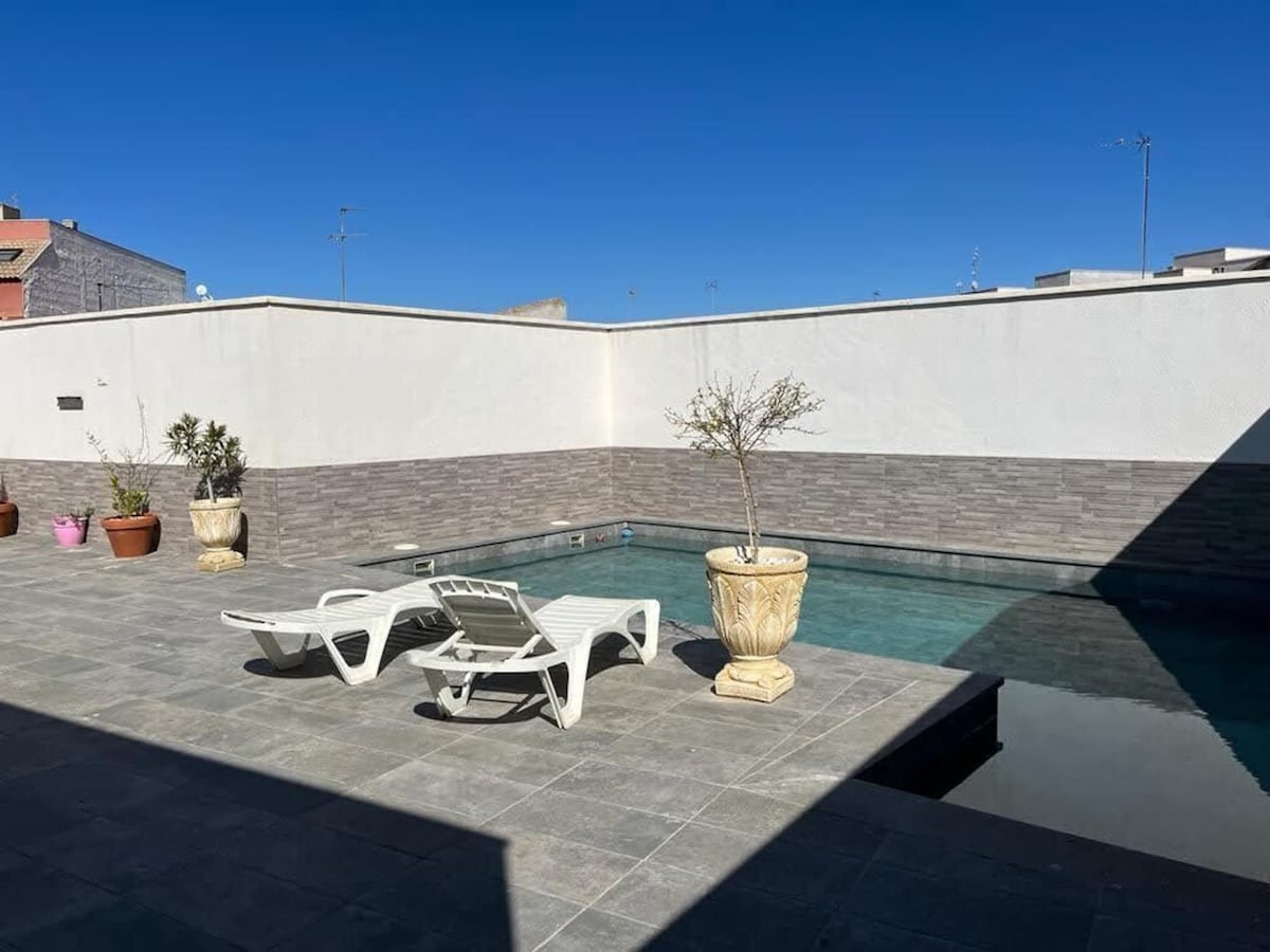 A private swimming pool is surrounded by sleek tiled flooring and clean white walls. Two lounge chairs are positioned beside the pool, while potted plants add touches of greenery to the area. Bright sunlight illuminates the serene space.
