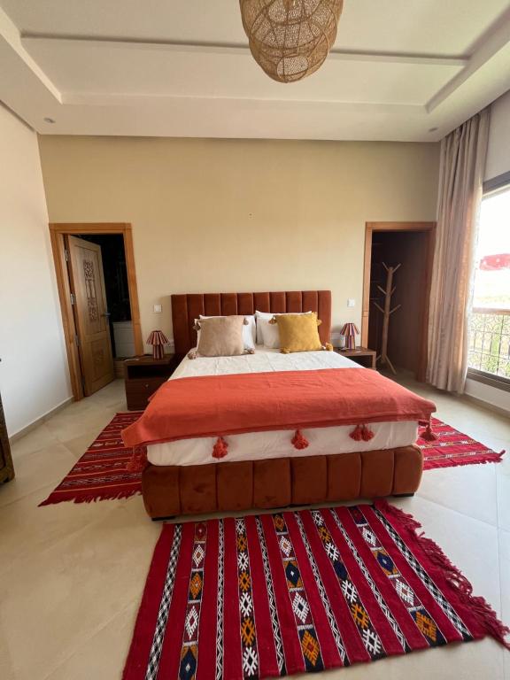 A well-appointed bedroom features a spacious bed with an orange throw and colorful decorative pillows. A richly patterned red rug lies on the floor, while large windows allow natural light to brighten the space. A wooden dresser and a coat rack complement the room's decor.