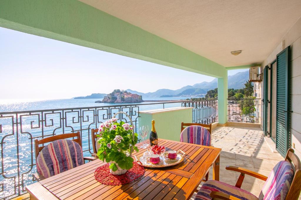 A spacious terrace features a wooden dining table surrounded by striped chairs. A vibrant arrangement of flowers is placed at the center, with sparkling water views visible beyond. The distant island and mountains create a serene backdrop under clear skies.