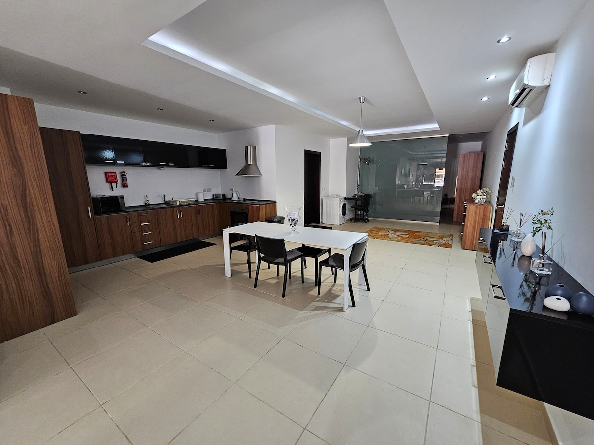 An open-plan living area is visible, featuring a modern kitchen with dark cabinetry and stainless steel appliances. A dining table surrounded by four black chairs occupies the center space. Light-colored tiles create a spacious ambiance, and additional furnishings add functionality to the area.
