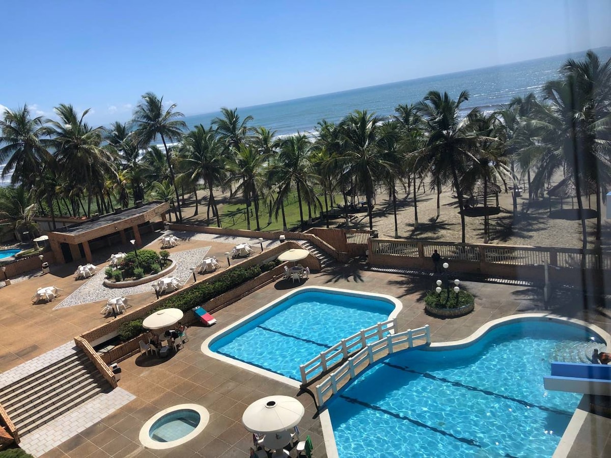 A panoramic view showcases a tranquil area featuring multiple swimming pools surrounded by palm trees. Sun loungers and umbrellas are arranged around the pools, while a pathway leads towards the beach in the distance, creating a serene coastal atmosphere.