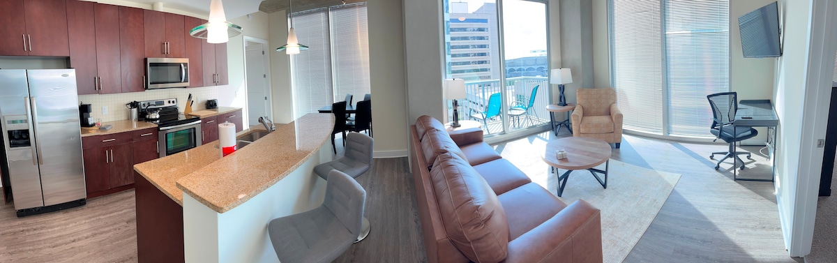 A bright and airy open-layout space features a fully equipped kitchen with modern appliances and a granite countertop. The living area includes comfortable seating, a coffee table, and a workspace. Floor-to-ceiling windows allow natural light to flood in, while a balcony with seating overlooks the city.