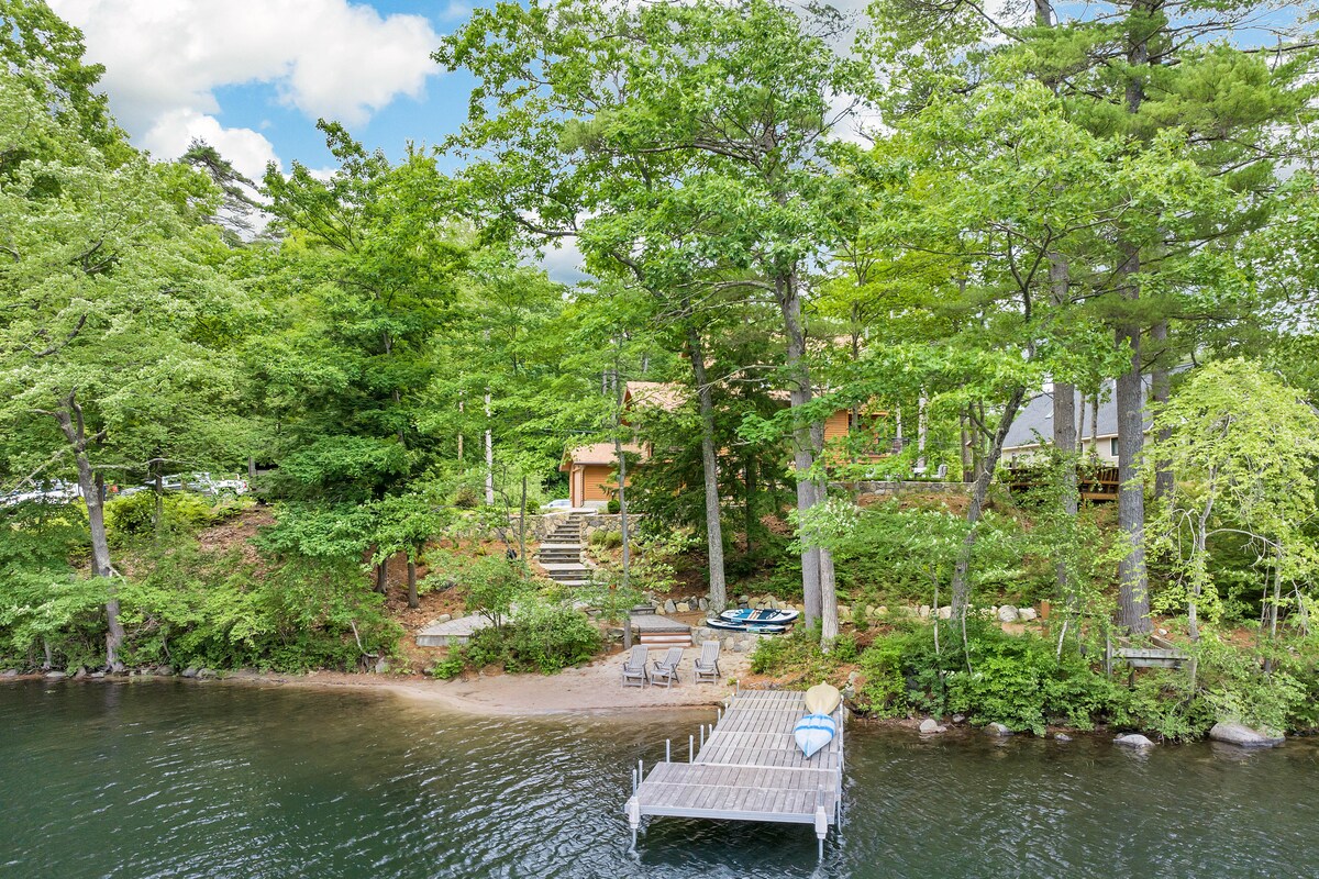 A serene waterfront scene showcases a private beach area framed by lush greenery. A wooden dock extends into the water, with several seating options available. Canoes and kayaks are visible on the shore, adjacent to a path leading to the home.