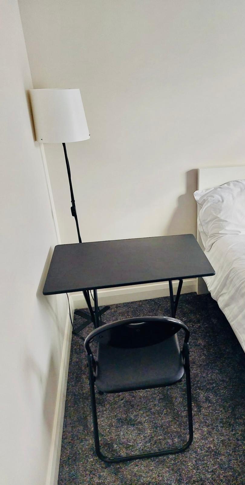 A compact workspace is provided with a black folding table and a chair. A floor lamp stands beside the table, casting light towards the sleeping area. The bed features a white duvet and is positioned against a light-colored wall, creating a minimalistic atmosphere.