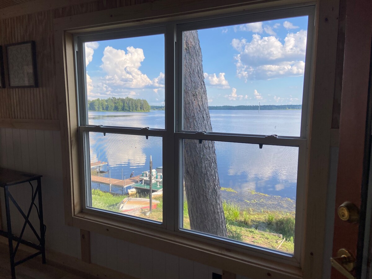 A large window frames a serene view of the St. Croix, displaying calm waters reflecting scattered clouds. Lush greenery and a private dock are visible in the foreground, providing a peaceful connection to the lake outside.