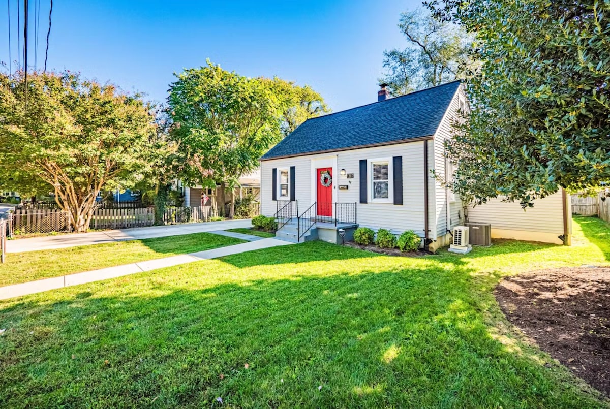 The exterior of a charming, single-story house is depicted, featuring a well-maintained lawn bordered by trees and shrubs. A vibrant red door provides a welcoming focal point. The pathway leads to the entrance, complemented by stairs on either side, indicating accessibility.