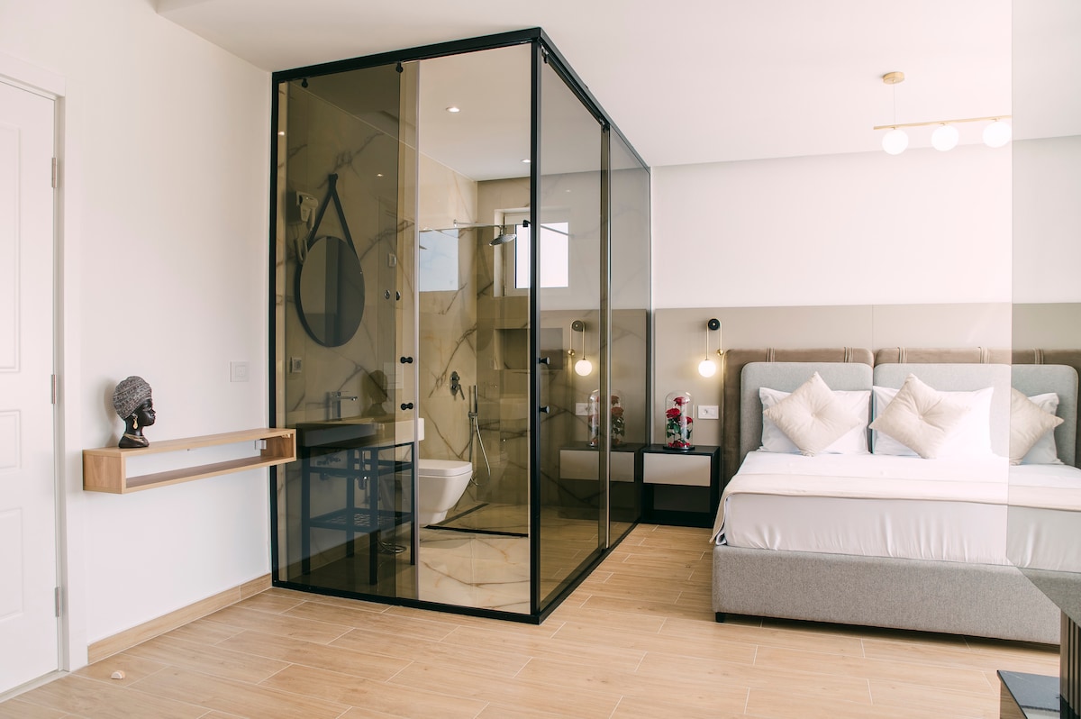 A spacious studio features a large bed with soft bedding, surrounded by neutral tones. The bathroom is separated by stylish glass walls. A small vanity area with a round mirror is visible, along with modern bedside tables holding ambient lighting.