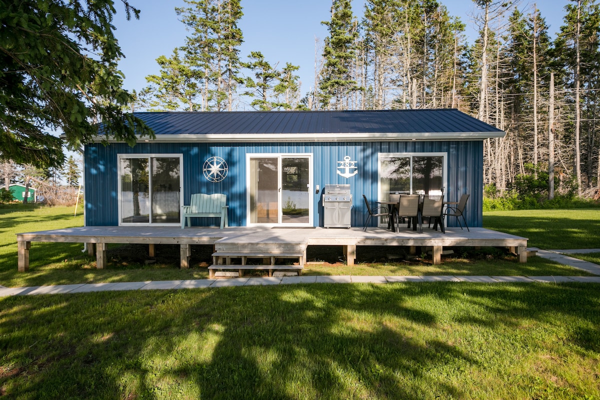 A modern blue cottage is presented, featuring large windows and a welcoming front porch. Outdoor seating and a grill are available, while a spacious green lawn enhances the setting. Tall trees provide a natural backdrop, complementing the cottage's inviting exterior.