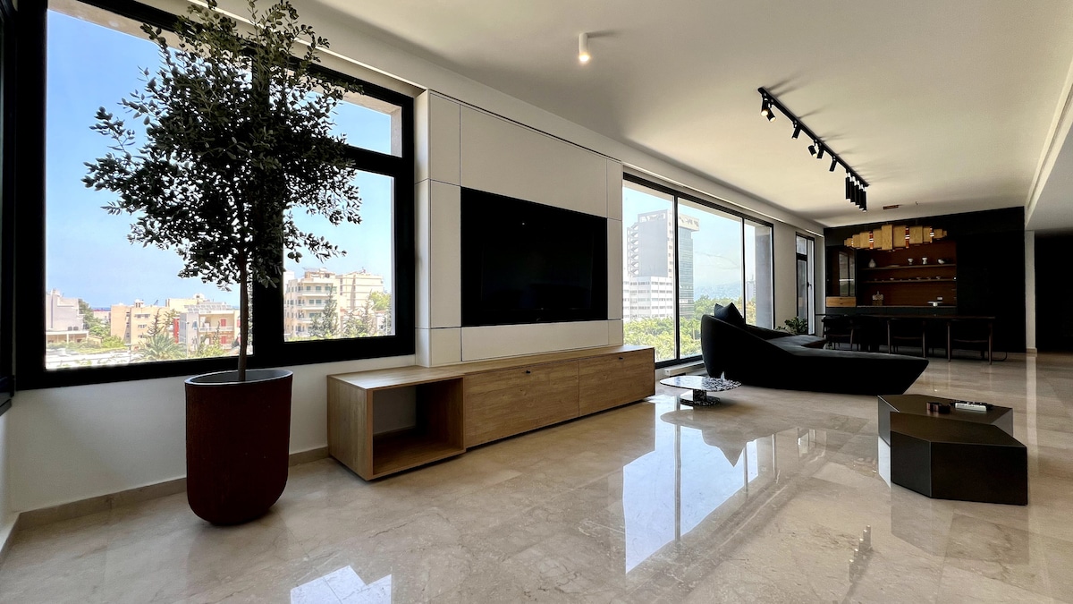 A stylish living area features large windows offering views of the surrounding cityscape. A sleek black sofa is positioned beside a light wood cabinet. A modern coffee table is situated on the polished stone floor, enhancing the spaciousness of the room.