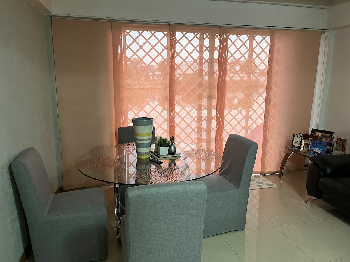 A dining area features a round glass table surrounded by four gray upholstered chairs. Soft, sheer curtains in a light hue provide a gentle filtering of light, while a decorative planter and small centerpiece enhance the table's simplicity. Framed photos are placed on a nearby surface.