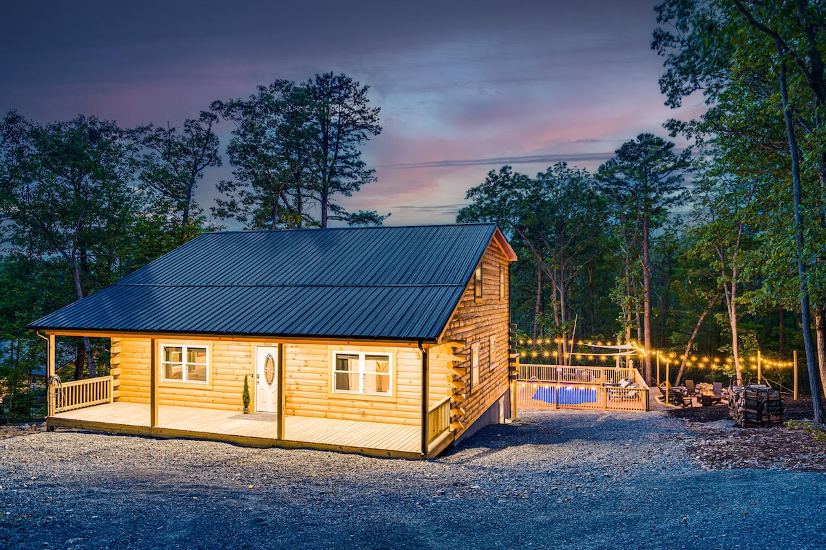 A classic log cabin design is nestled among the trees, with a warm glow from outdoor string lights illuminating the expansive deck. The serene setting features a gravel driveway and a welcoming entrance, while the scenic landscape provides a peaceful backdrop.