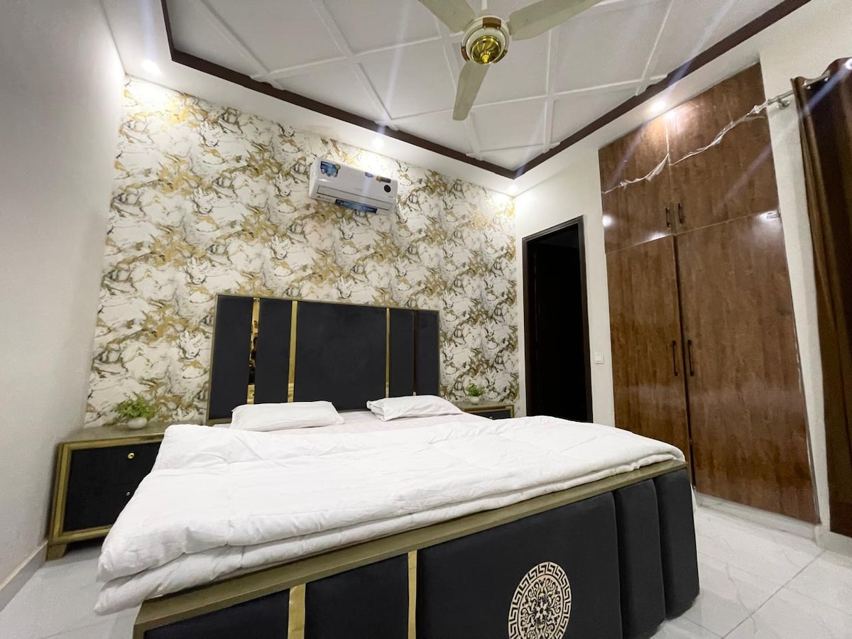 A spacious bedroom features a large bed with a decorative headboard, complemented by a stylish accent wall. The room is equipped with an air conditioning unit and includes a wardrobe for storage. Natural light fills the space, enhancing the modern decor.