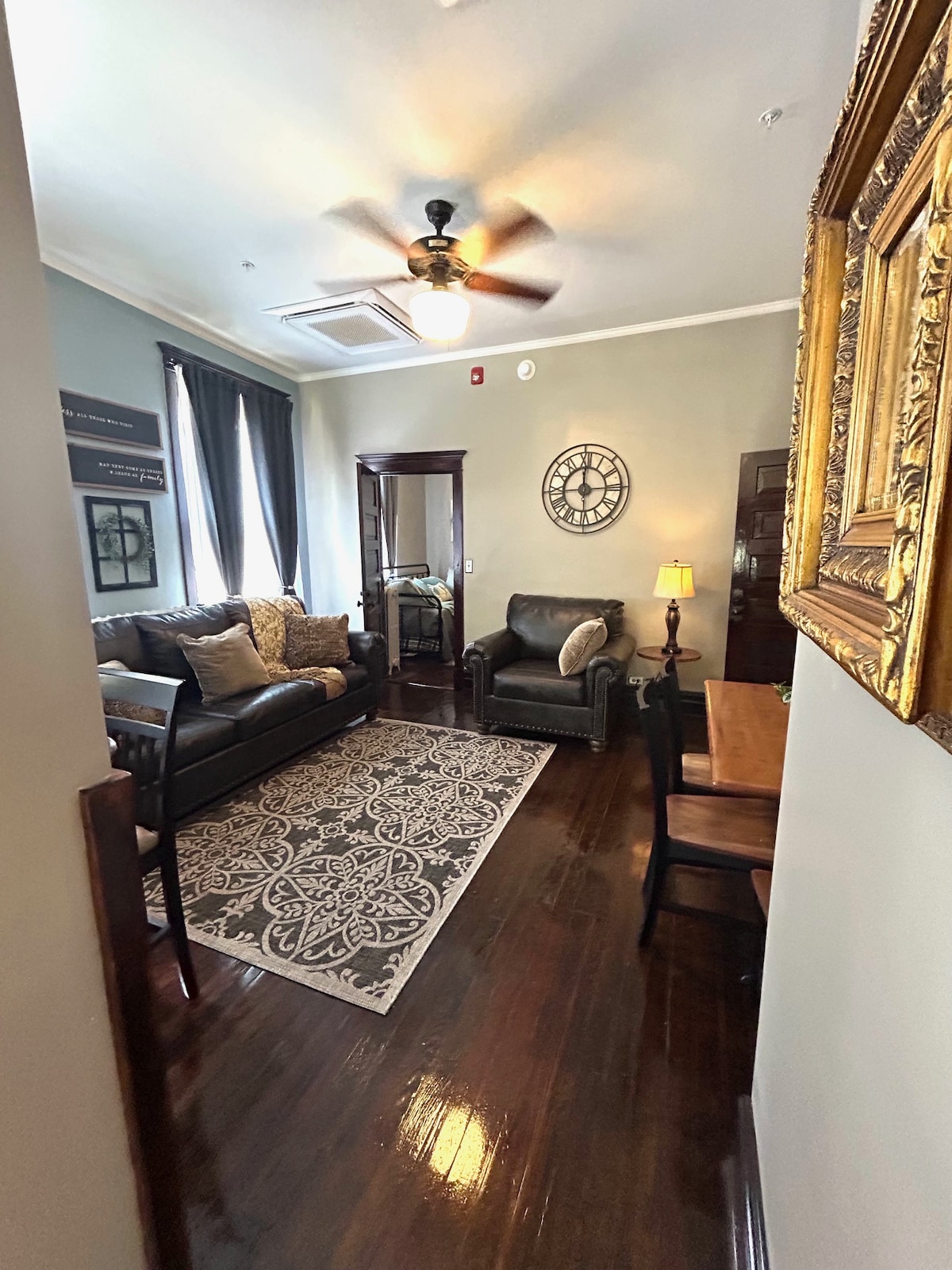A cozy living area features a dark leather sofa and a matching chair, complemented by soft throw pillows. A decorative rug lies beneath a small dining table, while a wall clock adds charm. Natural light filters through two tall windows, framed by dark curtains.