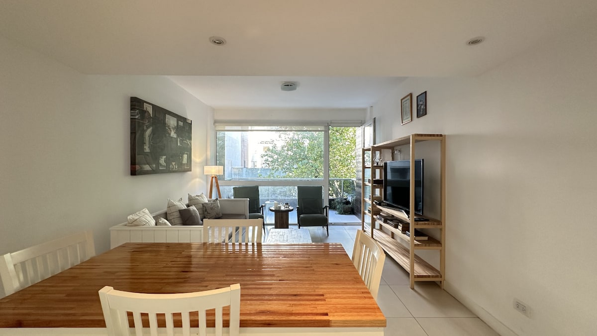 A bright living area features a light wooden dining table with white chairs. A comfortable seating arrangement includes two armchairs and a sofa, positioned near large windows that overlook greenery. A wooden shelving unit holds a television, enhancing the spacious feel of the open layout.
