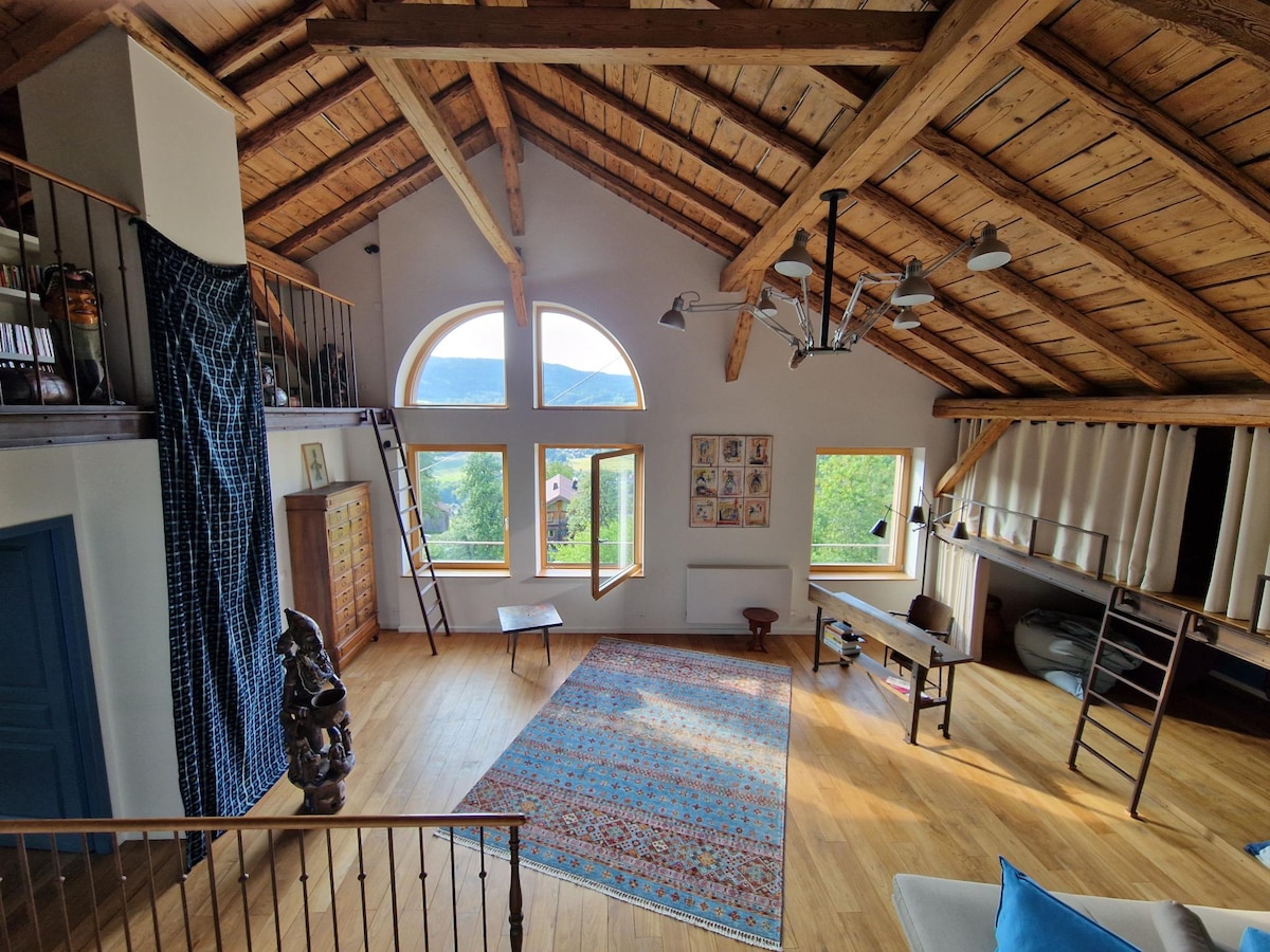 An open living area is highlighted by a cathedral ceiling with exposed wooden beams. Large windows offer ample natural light and views of the surrounding landscape. A colorful area rug lies on the hardwood floor, complementing the wooden furnishings and artistic decor adorning the walls.