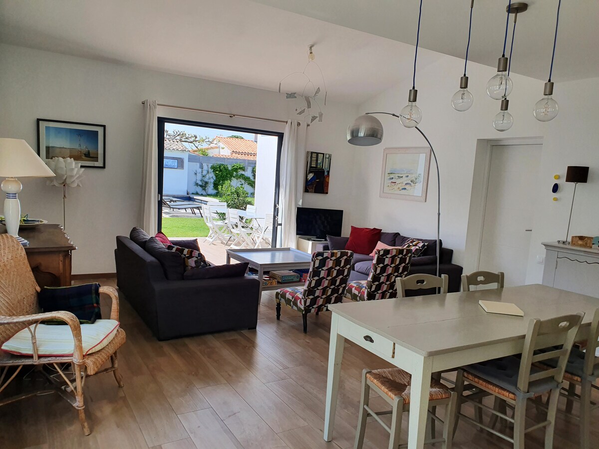 A bright living space features a mix of seating options, including a dark sofa and patterned chairs. A dining table is set nearby. Large glass doors open to a sunny courtyard, enhancing the connection between indoor and outdoor areas.