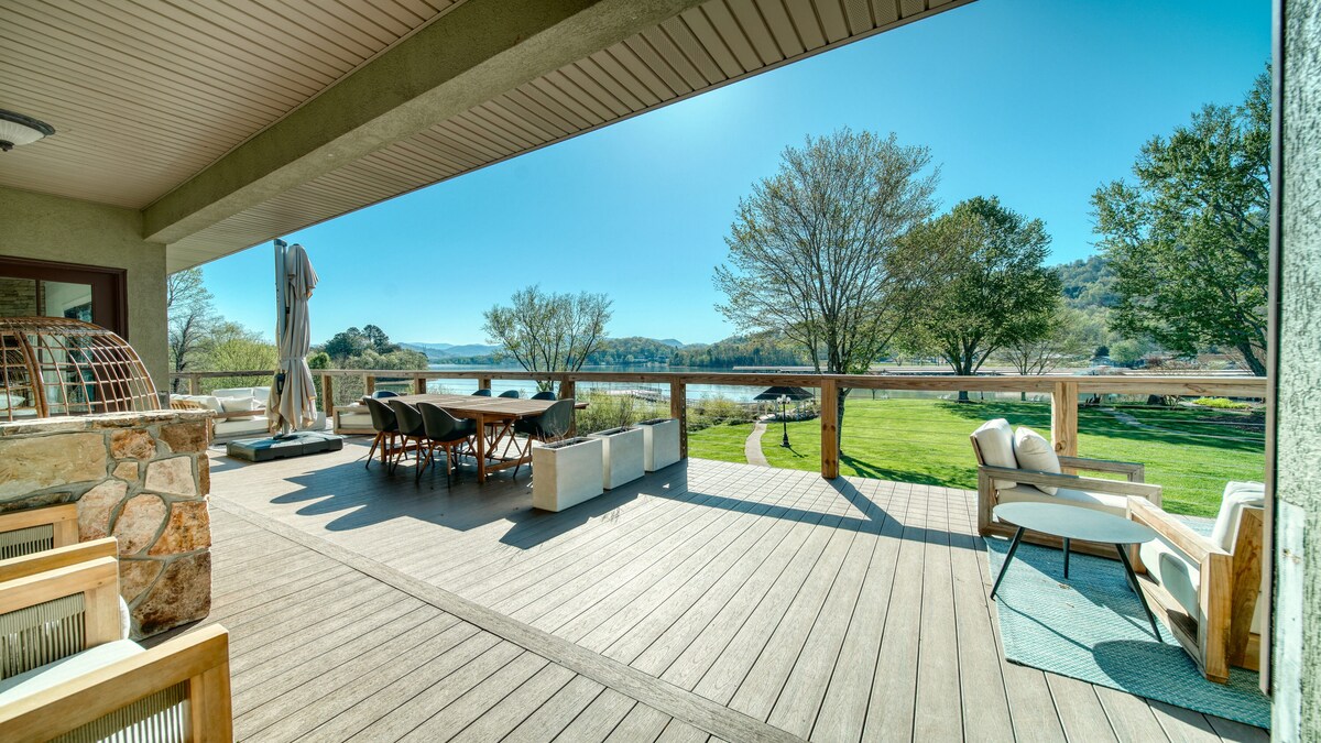 An outdoor patio area features a large dining table surrounded by chairs, offering views of a scenic landscape. Comfortable seating is arranged nearby, with sunlight illuminating the wooden deck. Lush greenery and trees are visible in the background, enhancing the natural ambiance.